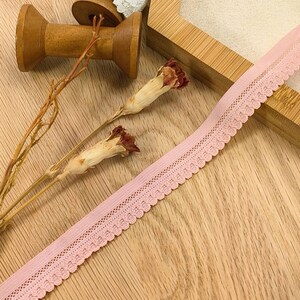 May include: Pink elastic lace trim with a scalloped edge. The trim is on a wooden surface with spools of thread and dried flowers.