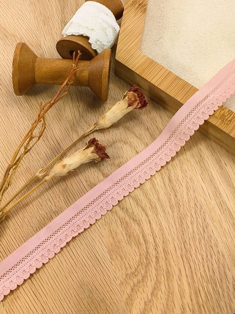 May include: Pink elastic lace trim with a scalloped edge. The trim is on a wooden surface with spools of thread and dried flowers.