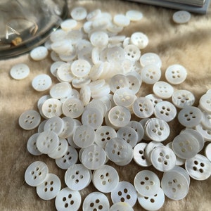 May include: A pile of white mother-of-pearl buttons with four holes each. The buttons are arranged in a random pattern on a light brown surface.