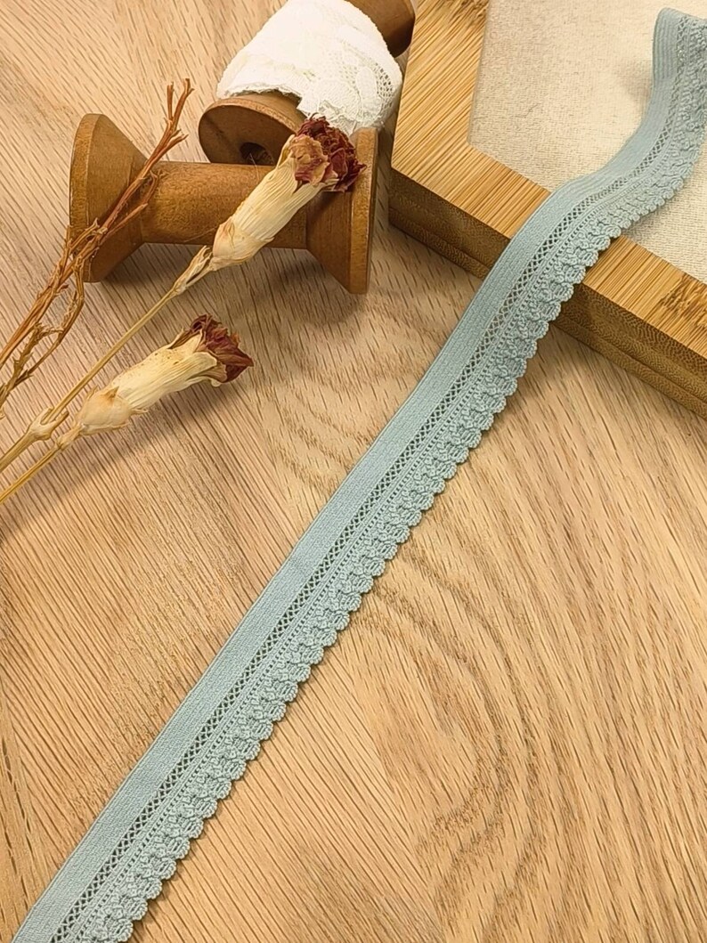 May include: A light blue elastic trim with a delicate lace pattern. The trim is shown on a wooden surface with spools of thread and dried flowers.
