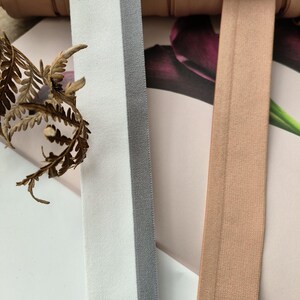 May include: Two rolls of elastic trim, one white with a grey stripe and one brown. The trim is lying on a striped fabric background.
