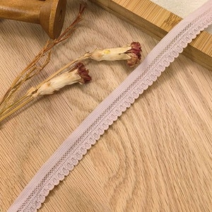 May include: A close-up of a pale pink elastic lace trim with a scalloped edge. The trim is lying on a wooden surface.
