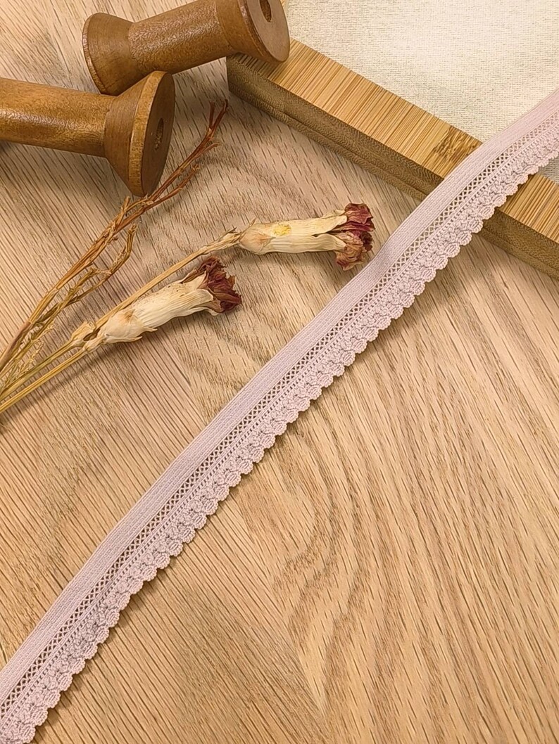 May include: A close-up of a pale pink elastic lace trim with a scalloped edge. The trim is lying on a wooden surface.