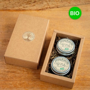 Könnte beinhalten: Zwei kleine, runde Metalldosen mit Deckeln, die Odd Polly's Deo Creme in einer braunen Pappbox enthalten. Die Dosen sind mit dem Text "Odd Polly's Deo Creme" und "BIO" beschriftet. Die Box ist mit einem Baumlogo verziert.