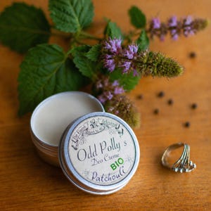 Op de afbeelding: Een blikje Odd Polly's Deo Creme Patchouli, met een gedeeltelijk open deksel, staat naast een ring en takjes met paarse bloemen en groene bladeren. Het etiket luidt "BIO". De achtergrond is een houten oppervlak.