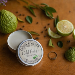 Op de afbeelding: Een open, ronde metalen pot van "Odd Polly's Deo Creme Bergamotte BIO" staat op een houten oppervlak, naast een ring en citrusvruchten. Het etiket van de pot is vintage-geïnspireerd. Groene citrusvruchten en bladeren zijn verspreid.