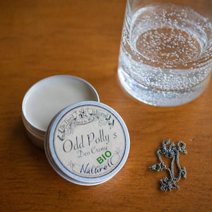 Könnte beinhalten: Eine offene, silberfarbene Dose Odd Polly's Deo Creme, mit dem Deckel daneben. Das Etikett lautet "BIO Naturell". Ein Glas Wasser und eine silberne Halskette befinden sich ebenfalls auf einer Holzoberfläche.