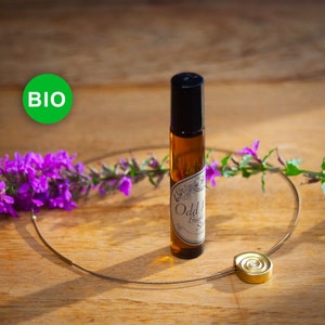 Könnte beinhalten: Eine braune Glasflasche mit einem schwarzen Roll-on-Verschluss und einem Etikett mit der Aufschrift "The Finest Things" und "Odd & Bright" mit einem floralen Design. Die Flasche steht auf einer Holzoberfläche mit einer goldenen Halskette mit einem runden Anhänger.