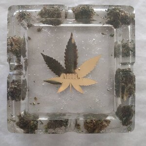 May include: A clear glass ashtray with a gold leaf design and the word "MOJO" inside. The ashtray is filled with small pieces of dried green plant material.