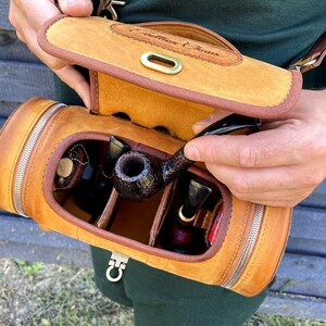 Puede incluir: Una funda de transporte de cuero marr&oacute;n para pipa con cierre de cremallera y una solapa con el texto "Southern Charms". La funda est&aacute; abierta y contiene varias pipas y una bolsa de tabaco.