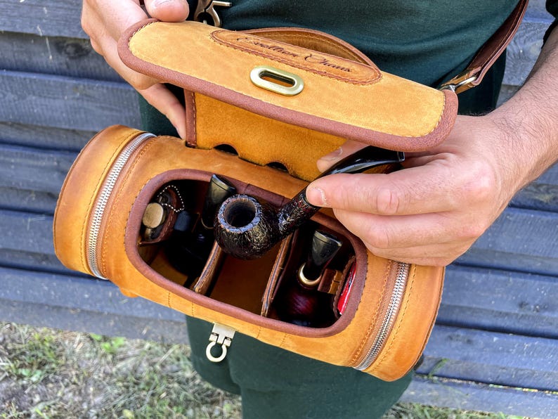 Puede incluir: Una funda de transporte de cuero marr&oacute;n para pipa con cierre de cremallera y una solapa con el texto "Southern Charms". La funda est&aacute; abierta y contiene varias pipas y una bolsa de tabaco.