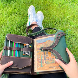 Puede incluir: Un cuaderno de bocetos de cuero marr&oacute;n con una solapa de cuero verde y un bolsillo con cremallera para l&aacute;pices. El cuaderno est&aacute; abierto para revelar una p&aacute;gina en blanco y una p&aacute;gina con una ilustraci&oacute;n colorida de un edificio. El texto "Alexandra" y "College Block" es visible en el cuaderno.