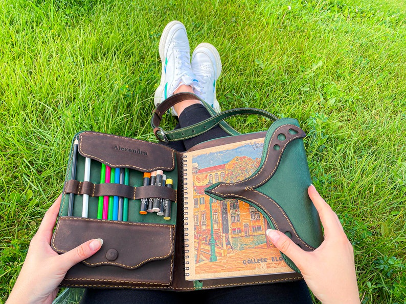 Puede incluir: Un cuaderno de bocetos de cuero marr&oacute;n con una solapa de cuero verde y un bolsillo con cremallera para l&aacute;pices. El cuaderno est&aacute; abierto para revelar una p&aacute;gina en blanco y una p&aacute;gina con una ilustraci&oacute;n colorida de un edificio. El texto "Alexandra" y "College Block" es visible en el cuaderno.