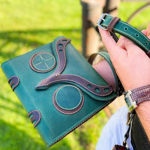 Puede incluir: Un diario de cuero verde con una correa de cuero marr&oacute;n y costuras decorativas. El diario tiene un dise&ntilde;o &uacute;nico con un patr&oacute;n circular y una correa en forma de Y.