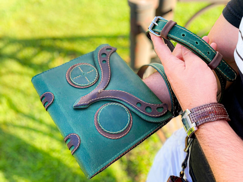 Puede incluir: Un diario de cuero verde con una correa de cuero marr&oacute;n y costuras decorativas. El diario tiene un dise&ntilde;o &uacute;nico con un patr&oacute;n circular y una correa en forma de Y.