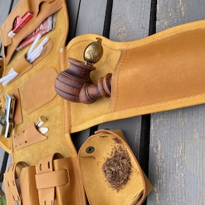 Puede incluir: Un estuche de cuero marrón claro para pipa de tabaco, abierto para mostrar su contenido. Incluye una pipa de madera, tabaco, herramientas de limpieza y un encendedor. El estuche se muestra sobre una superficie de madera.