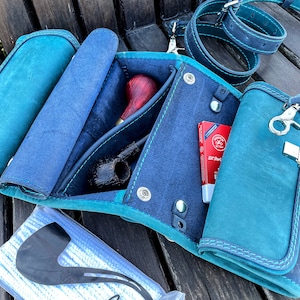 May include: A teal leather pipe tobacco pouch with a blue lining. The pouch has a flap that opens to reveal a pipe, a pack of pipe filters, and a small pouch for tobacco. The pouch has a strap and a metal clasp.