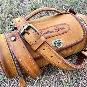 Puede incluir: Una bolsa cil&iacute;ndrica de cuero marr&oacute;n con una correa y un cierre de cremallera. La bolsa tiene un asa de cuero marr&oacute;n y una hebilla de metal. La bolsa est&aacute; estampada con el texto "Evolution Chaos".