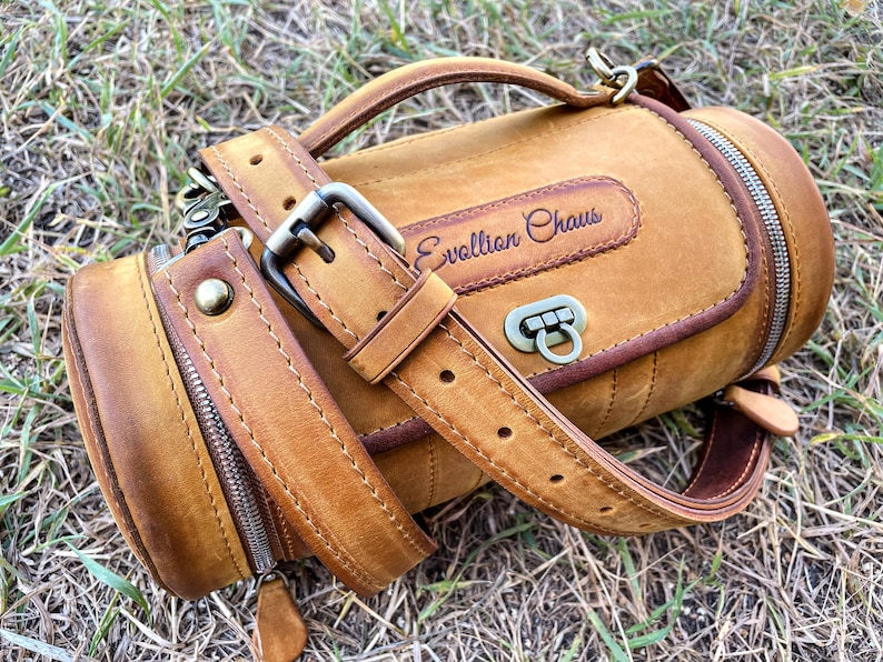 Puede incluir: Una bolsa cil&iacute;ndrica de cuero marr&oacute;n con una correa y un cierre de cremallera. La bolsa tiene un asa de cuero marr&oacute;n y una hebilla de metal. La bolsa est&aacute; estampada con el texto "Evolution Chaos".