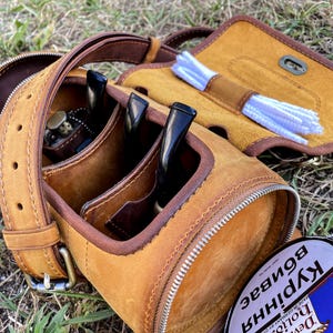 Puede incluir: Una funda de transporte de cuero marr&oacute;n para pipa con cierre de cremallera. La funda tiene tres ranuras para pipas y un compartimento separado para limpiadores de pipas. La funda est&aacute; sentada sobre un lecho de hierba verde.