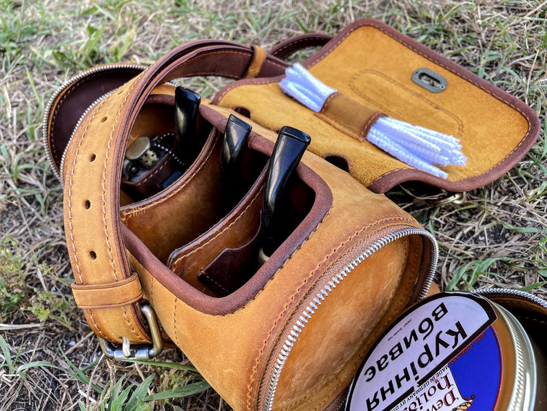 Puede incluir: Una funda de transporte de cuero marr&oacute;n para pipa con cierre de cremallera. La funda tiene tres ranuras para pipas y un compartimento separado para limpiadores de pipas. La funda est&aacute; sentada sobre un lecho de hierba verde.