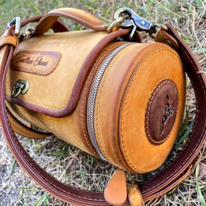 Puede incluir: Una bolsa cil&iacute;ndrica de cuero marr&oacute;n con cierre de cremallera y una correa. La bolsa tiene una solapa de cuero marr&oacute;n con el texto "Cowhide Craft" grabado.