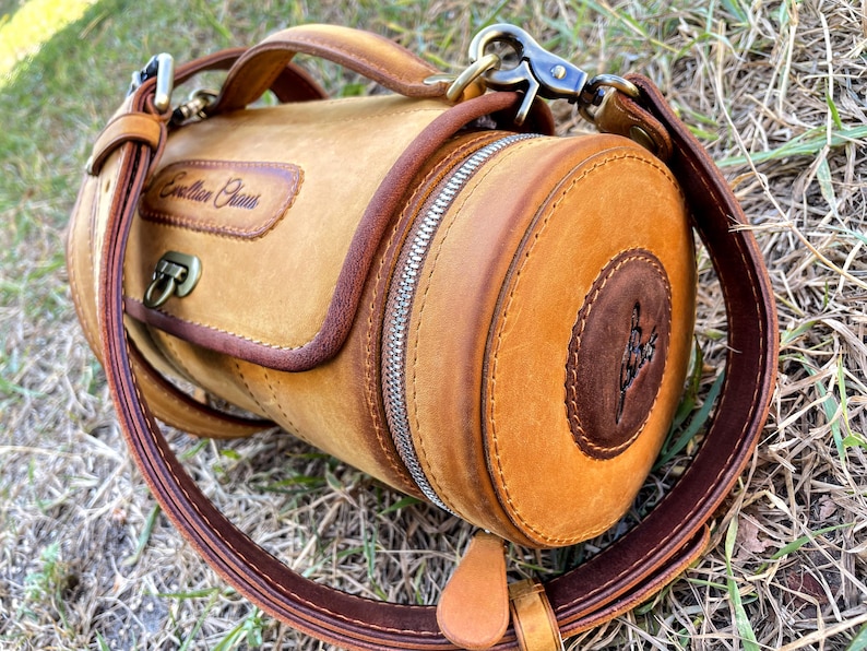 Puede incluir: Una bolsa cil&iacute;ndrica de cuero marr&oacute;n con cierre de cremallera y una correa. La bolsa tiene una solapa de cuero marr&oacute;n con el texto "Cowhide Craft" grabado.