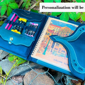 May include: A teal blue leather sketchbook with a flap that has a pocket for pens and pencils. The sketchbook is open to reveal a blank page with the text "COLLEGE BLO" printed on it. The sketchbook is sitting on a bed of rocks and green foliage.