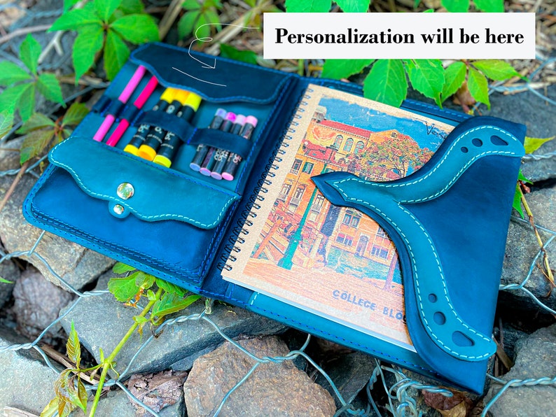 May include: A teal blue leather sketchbook with a flap that has a pocket for pens and pencils. The sketchbook is open to reveal a blank page with the text "COLLEGE BLO" printed on it. The sketchbook is sitting on a bed of rocks and green foliage.
