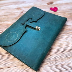 May include: A teal leather notebook with a decorative clasp in the shape of a violin. The notebook is lying on a wooden surface.