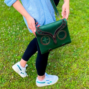Puede incluir: Un bolso de cuero verde cruzado con un dise&ntilde;o de b&uacute;ho. El bolso tiene detalles y costuras de cuero marr&oacute;n. El bolso lo lleva una persona que lleva pantalones negros y zapatillas blancas.