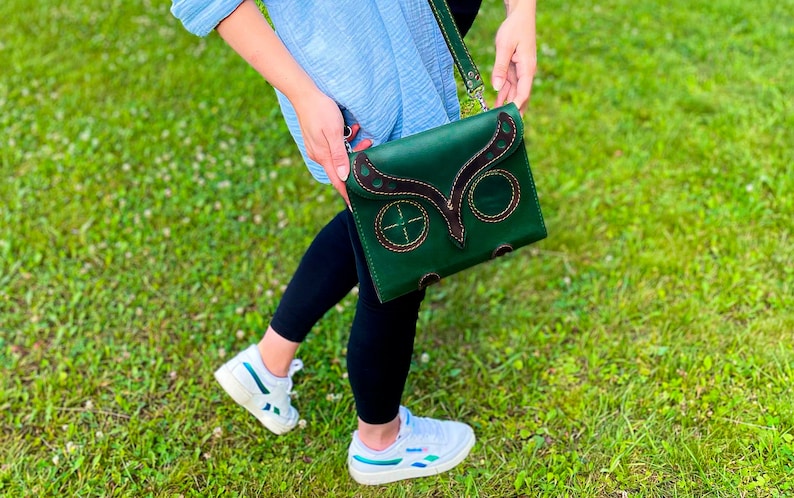 Puede incluir: Un bolso de cuero verde cruzado con un dise&ntilde;o de b&uacute;ho. El bolso tiene detalles y costuras de cuero marr&oacute;n. El bolso lo lleva una persona que lleva pantalones negros y zapatillas blancas.