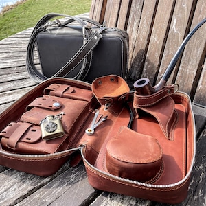 Puede incluir: Un estuche de viaje de cuero marrón con múltiples compartimentos, incluyendo un bolsillo para un encendedor, una pipa y una pequeña bolsa. El estuche está abierto y el interior es visible. El estuche está sentado sobre una superficie de madera.