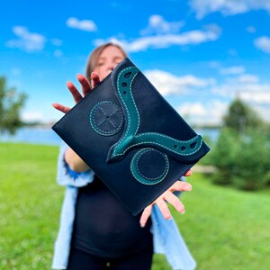 May include: A black leather clutch with a green leather design. The design features a stylised bird with a circular pattern on the front.