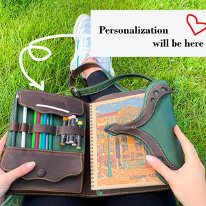 Puede incluir: Un cuaderno de cuero marr&oacute;n con una solapa de cuero verde y un cierre de cremallera. El cuaderno tiene un porta l&aacute;pices incorporado y un espacio para la personalizaci&oacute;n. El cuaderno est&aacute; abierto para revelar una p&aacute;gina en blanco con una ilustraci&oacute;n colorida de un edificio.