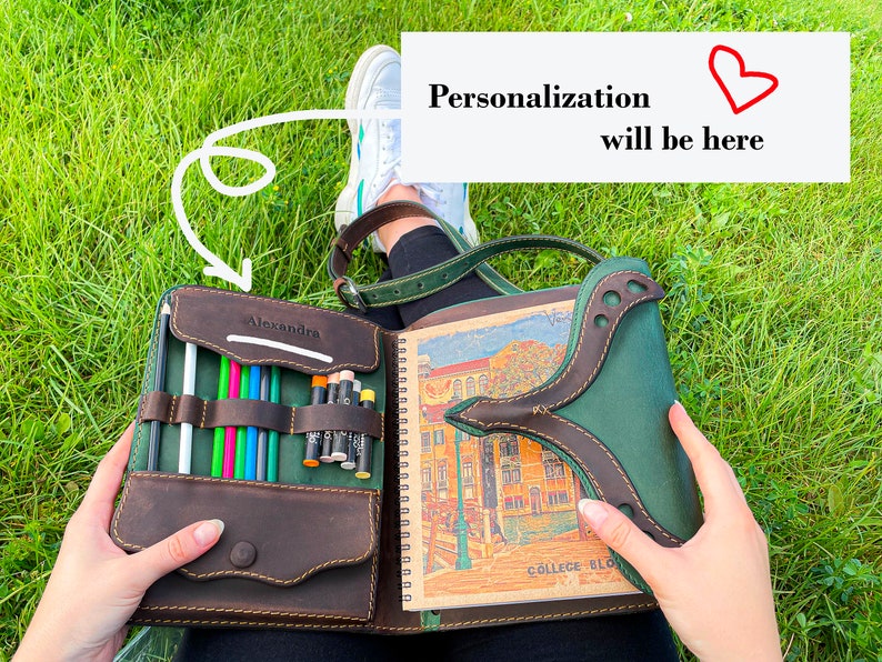 Puede incluir: Un cuaderno de cuero marr&oacute;n con una solapa de cuero verde y un cierre de cremallera. El cuaderno tiene un porta l&aacute;pices incorporado y un espacio para la personalizaci&oacute;n. El cuaderno est&aacute; abierto para revelar una p&aacute;gina en blanco con una ilustraci&oacute;n colorida de un edificio.
