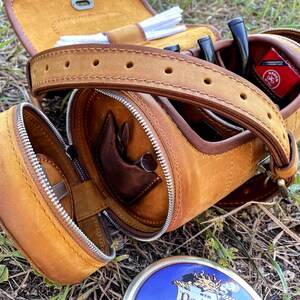 Puede incluir: Una bolsa de tabaco de cuero marr&oacute;n con cierre de cremallera y una correa. La bolsa tiene varios compartimentos para guardar tabaco, pipas y otros accesorios. La bolsa est&aacute; abierta y el interior es visible.