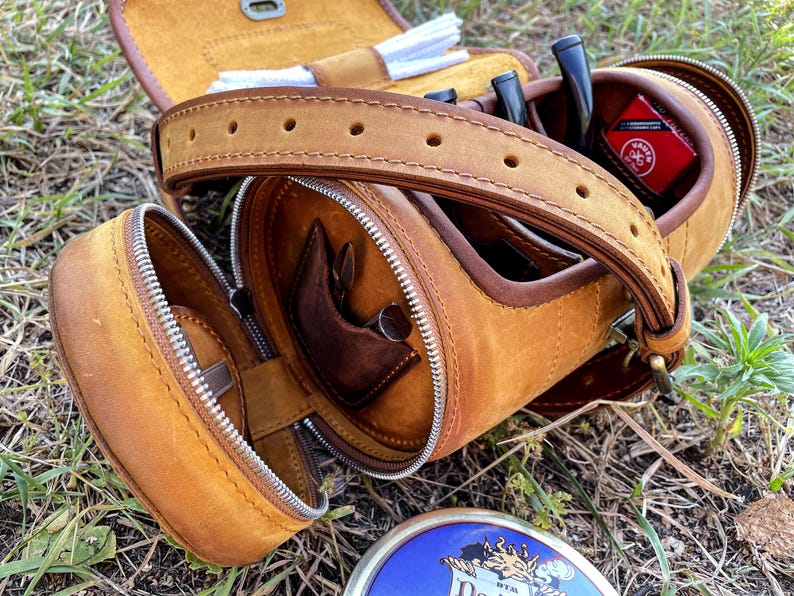 Puede incluir: Una bolsa de tabaco de cuero marr&oacute;n con cierre de cremallera y una correa. La bolsa tiene varios compartimentos para guardar tabaco, pipas y otros accesorios. La bolsa est&aacute; abierta y el interior es visible.