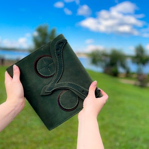 May include: A dark green leather journal with decorative stitching and circular cutouts. The journal is being held in someone's hands.