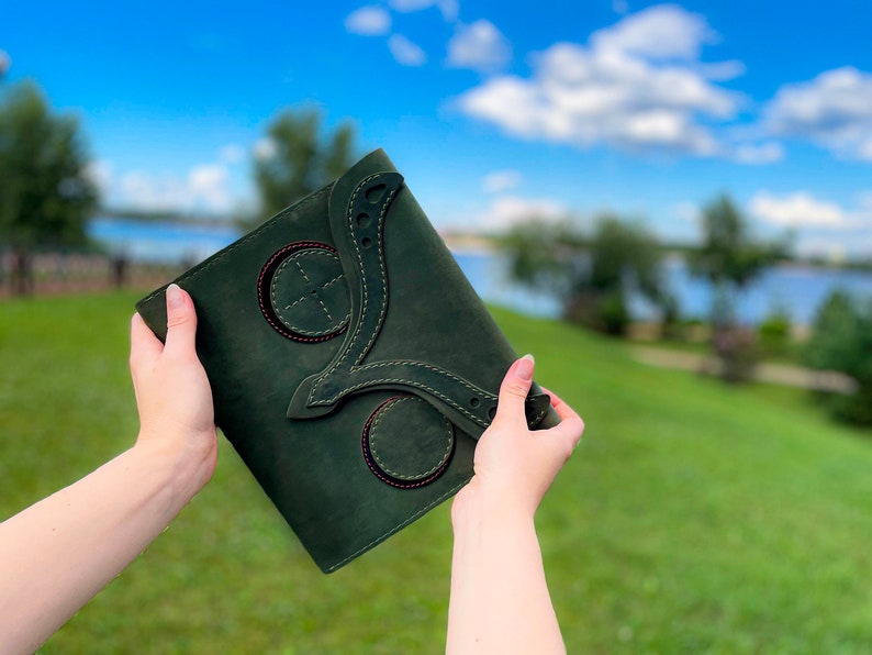 May include: A dark green leather journal with decorative stitching and circular cutouts. The journal is being held in someone's hands.