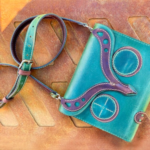 May include: A teal leather crossbody bag with brown leather accents and decorative stitching. The bag has a flap closure and a long adjustable strap.