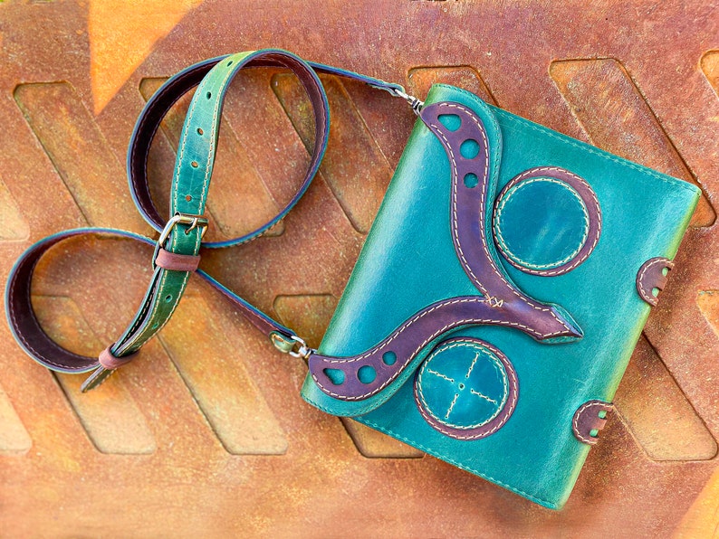 Puede incluir: Un bolso bandolera de cuero turquesa con detalles en cuero marr&oacute;n y costuras decorativas. El bolso tiene cierre de solapa y una correa larga ajustable.