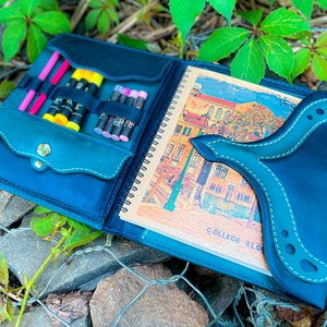 May include: A blue leather sketchbook with a flap closure and a built-in pencil holder. The sketchbook has a spiral binding and a colourful illustration on the inside. The text "COLLEGE BLO" is visible on the inside page.