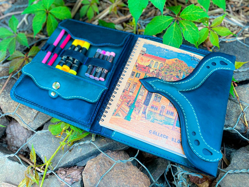 May include: A blue leather sketchbook with a flap closure and a built-in pencil holder. The sketchbook has a spiral binding and a colourful illustration on the inside. The text "COLLEGE BLO" is visible on the inside page.