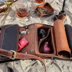 May include: A brown leather tobacco pipe roll with a shoulder strap, open to reveal a pipe, tobacco, and accessories. A bottle of liquor and a glass of amber liquid are in the background. The roll is on a sandy surface.