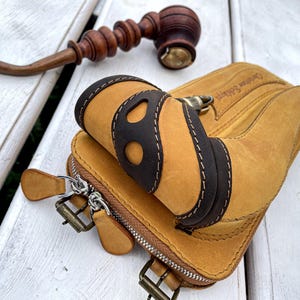 May include: A tan leather glove with dark brown accents and white stitching, featuring a zipper closure and metal hardware. A wooden pipe and mallet are in the background. The glove has the text "Genuine Selberg" on it.