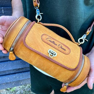 Puede incluir: Una bolsa cil&iacute;ndrica de cuero marr&oacute;n con cierre de cremallera y una correa. La bolsa tiene una hebilla de lat&oacute;n y el texto "Evolution Chaus" est&aacute; grabado en la parte delantera.