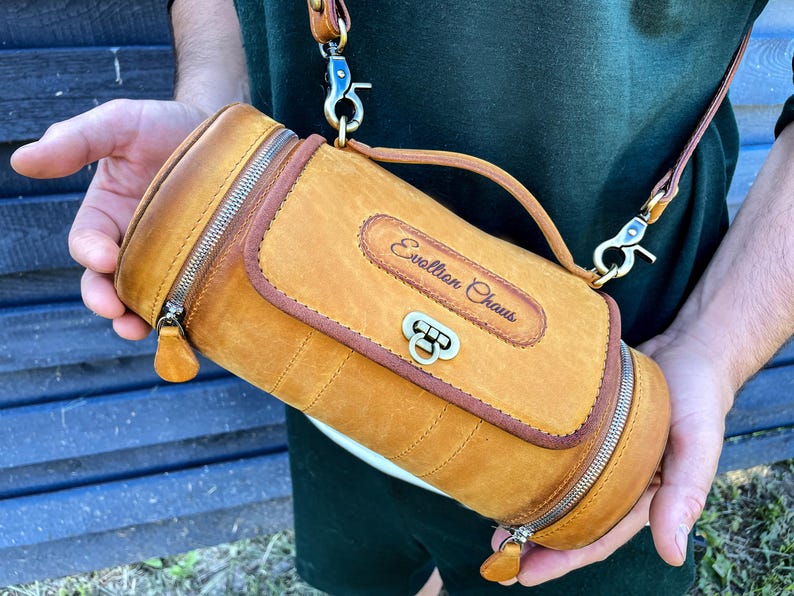 Puede incluir: Una bolsa cil&iacute;ndrica de cuero marr&oacute;n con cierre de cremallera y una correa. La bolsa tiene una hebilla de lat&oacute;n y el texto "Evolution Chaus" est&aacute; grabado en la parte delantera.