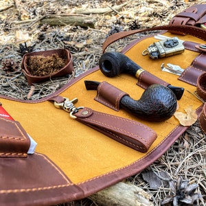 May include: A brown leather tobacco pouch with a yellow suede lining. The pouch has multiple pockets for storing pipes, a lighter, and other accessories. The pouch is unzipped and laid flat on a bed of pine needles.