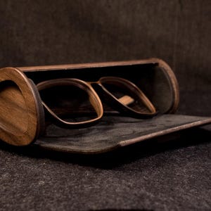 Könnte beinhalten: Brille und Etui aus Holz. Die Brille hat einen dunkelbraunen Rahmen und liegt in einem passenden Holz-Etui. Das Etui ist geöffnet und gibt die Brille frei. Der Hintergrund ist eine dunkle, strukturierte Oberfläche.